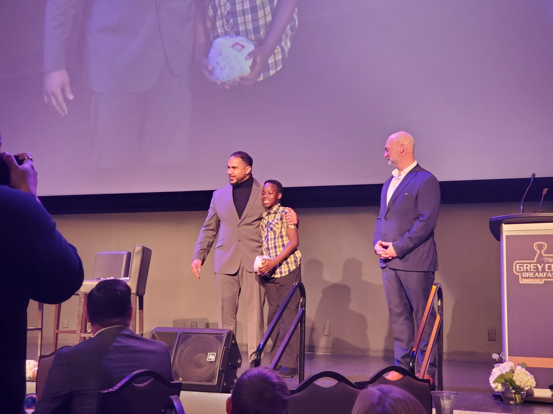 A young boy stands on stage holding a football and is flanked by 2 men
